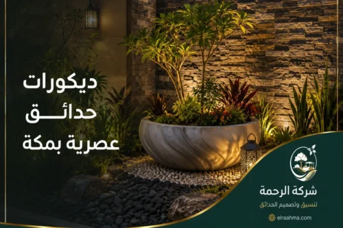 زراعة وتنسيق حدائق بمكة - شركة الرحمة تنسيق حوض نباتات بمكة أمام جدار حجري مع أشجار ونباتات متنوعة وإضاءة خارجية.